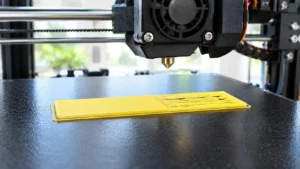 Teste de primeira camada em PLA amarelo-claro sobre chapa PEI preta, mostrando linhas corretamente unidas e outra região com lacunas entre trajetórias, com bico da impressora 3D estacionado acima da peça.